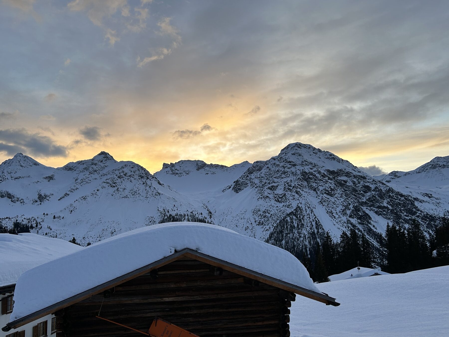 Abendrot über den verschneiten Gipfeln, davor ein Dach unter der Schneelast
