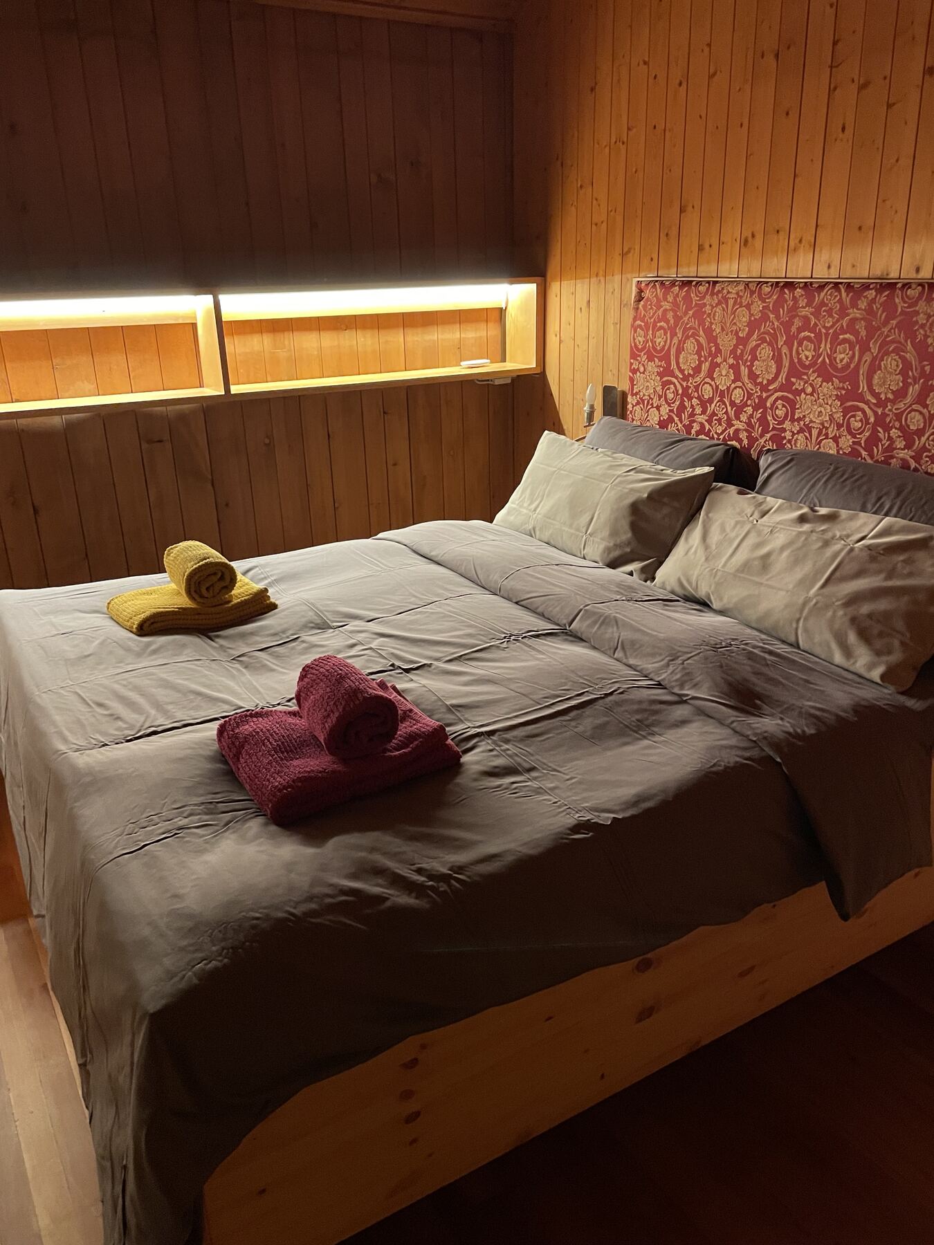 Das Elternzimmer mit Doppelbett vor der Holzwand, abends bei indirektem Licht